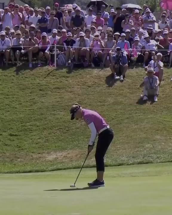 A golfer in a pink and white top and black pants putts on a green while a large gallery watches from bleachers along a hillside, then celebrates with a fist pump while walking off the green next to her caddie in a pink bib.