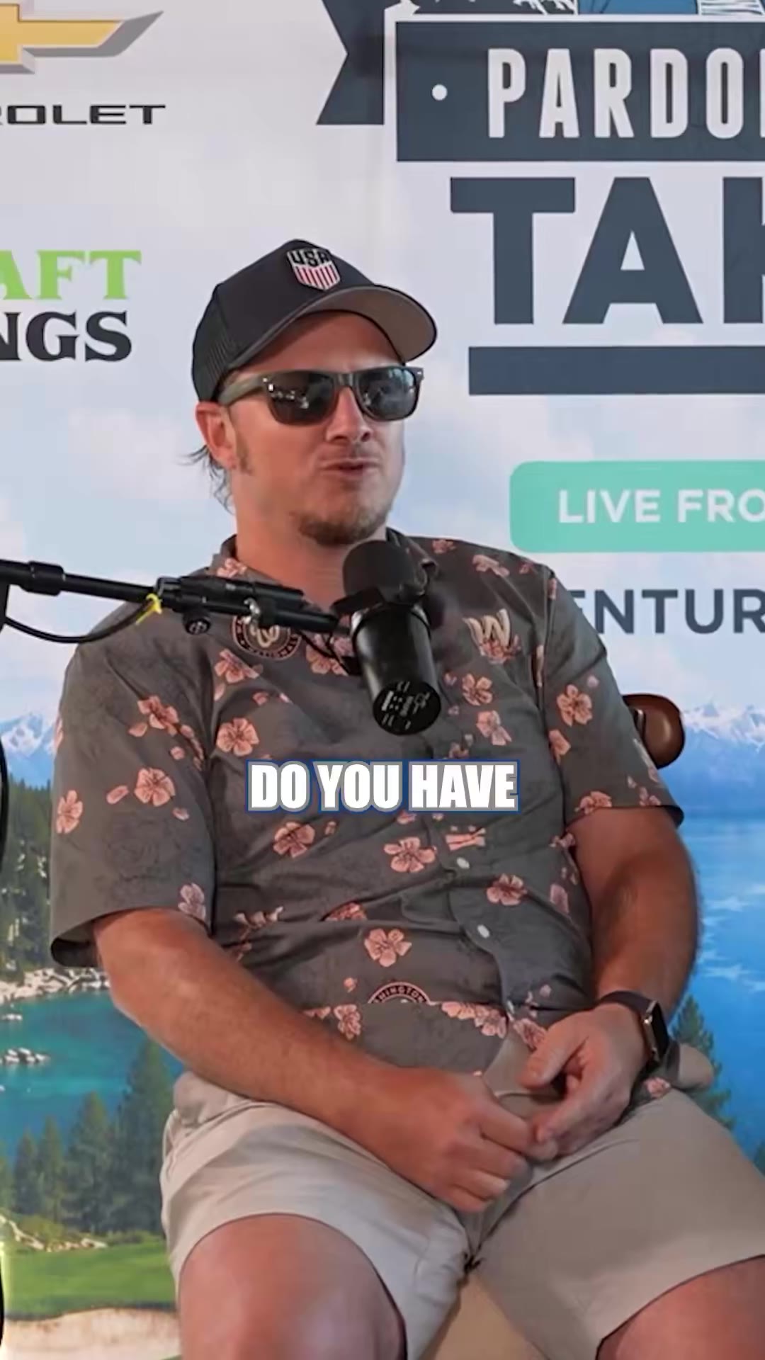 A man wearing a white cap and dark polo shirt sits in a chair speaking into a podcast microphone in front of a 'Pardon My Take' backdrop featuring lake and mountain scenery, gesturing with his hands while talking and smiling.