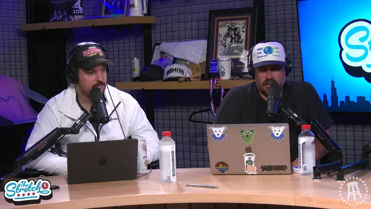 Two podcast hosts sit at a desk with laptops, water bottles, and BodyArmor drinks in front of a 'The Stretch' branded backdrop featuring a Chicago skyline graphic, talking and gesturing during recording.
