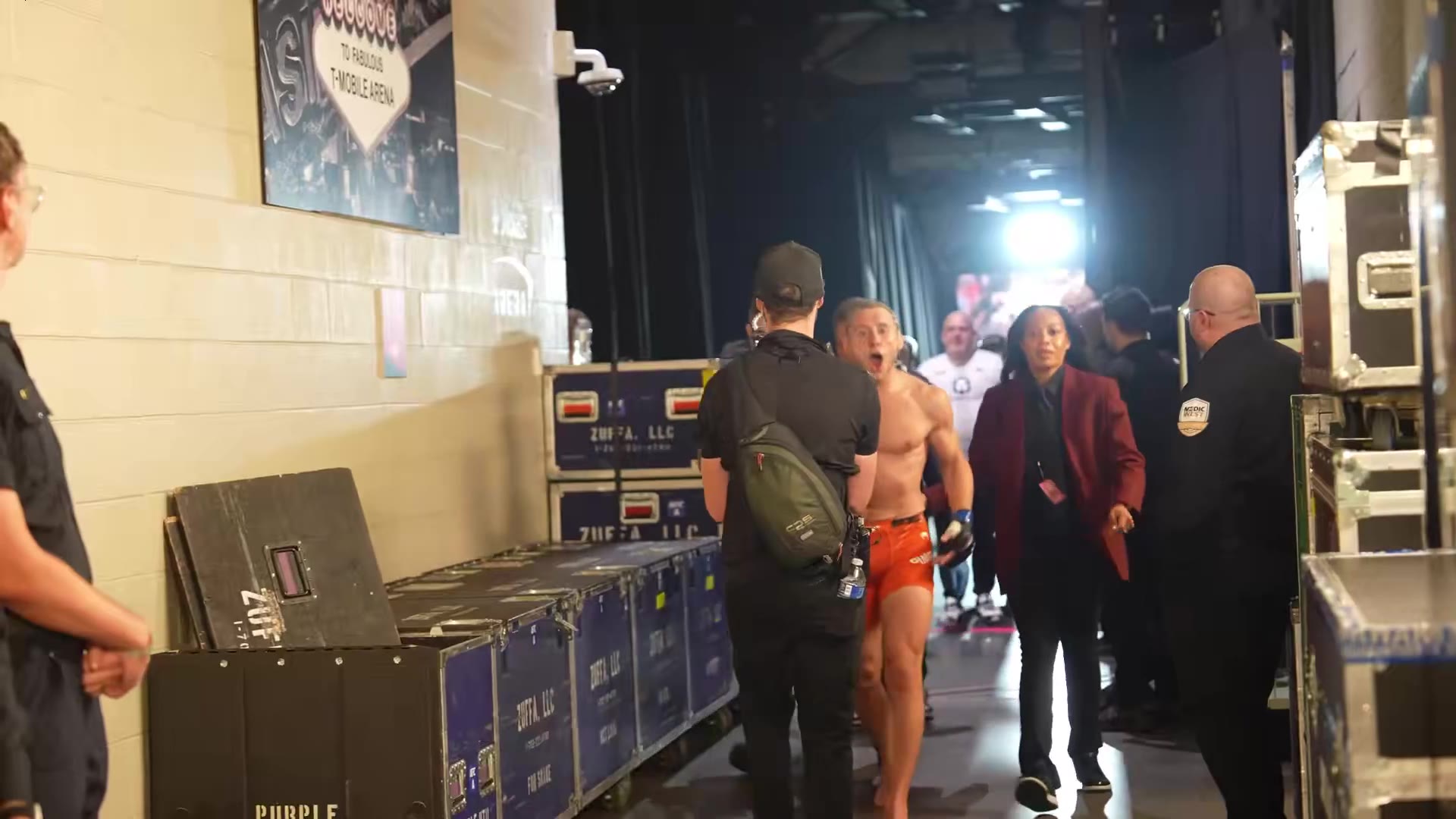 A man showers under bright light then walks through a backstage/arena area lit with blue and purple lighting, talking to the camera, before biting into a slice of pepperoni pizza in a hallway near other people, with a partially visible sign reading 'obile'.