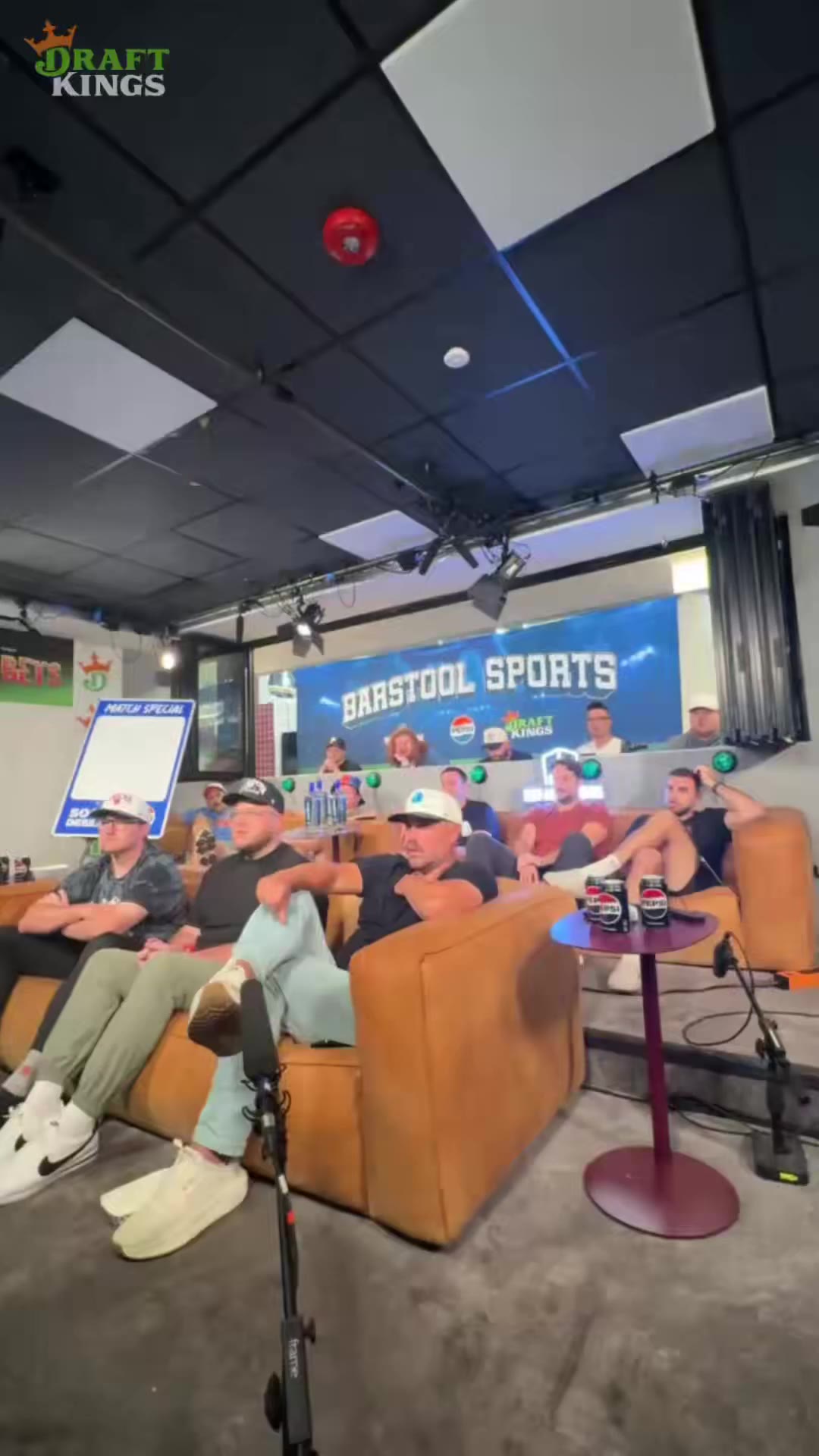 Group of men in casual clothes and caps sit on tan leather couches on a podcast/broadcast set with a large screen behind them reading BARSTOOL SPORTS, watching something off-screen with excited reactions and raised arms.