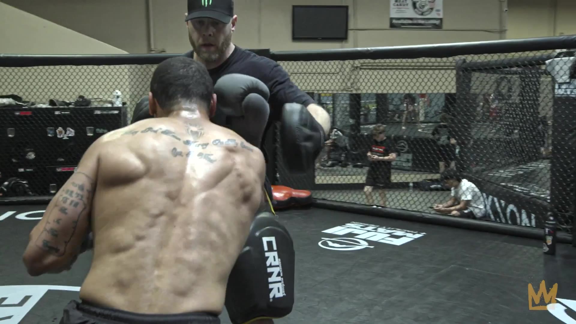 Two men sit on the mat inside a fenced MMA cage, one shirtless and one wearing a black shirt with a Celtics logo, then cuts to a close-up of the man in the black Celtics shirt speaking as he sits against the cage.