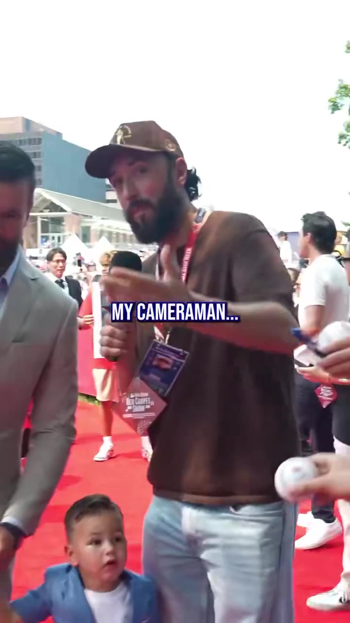 Two men on a red carpet event, one wearing a brown t-shirt with a lanyard and cap holding a microphone with a network logo, talking to other men in suits; on-screen captions read EXACTLY and NO? EXACTLY.