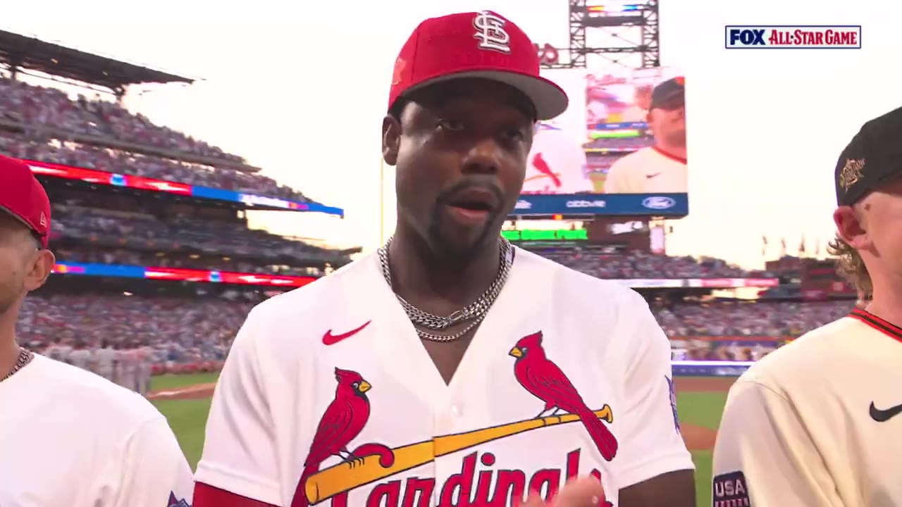 Jordan Walker, wearing a red St. Louis Cardinals All-Star jersey and cap with a silver chain necklace, smiles and waves to the crowd on the field at an MLB All-Star Game, with a fan in the stands wearing a Cardinals shirt applauding in the crowd.