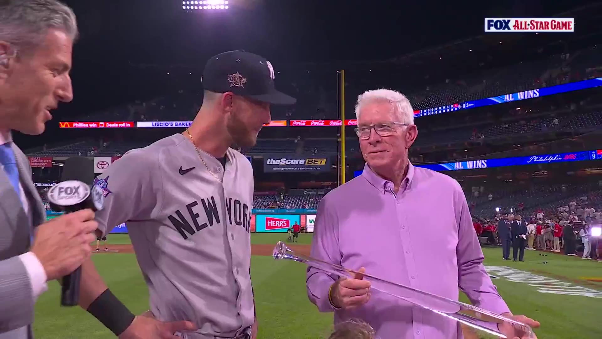 A sideline reporter interviews a New York-uniformed baseball player holding a trophy shaped like a sword/dagger at a stadium during the All-Star Game; a young girl in a pink shirt stands nearby, with an older man in a lavender shirt laughing in the background.