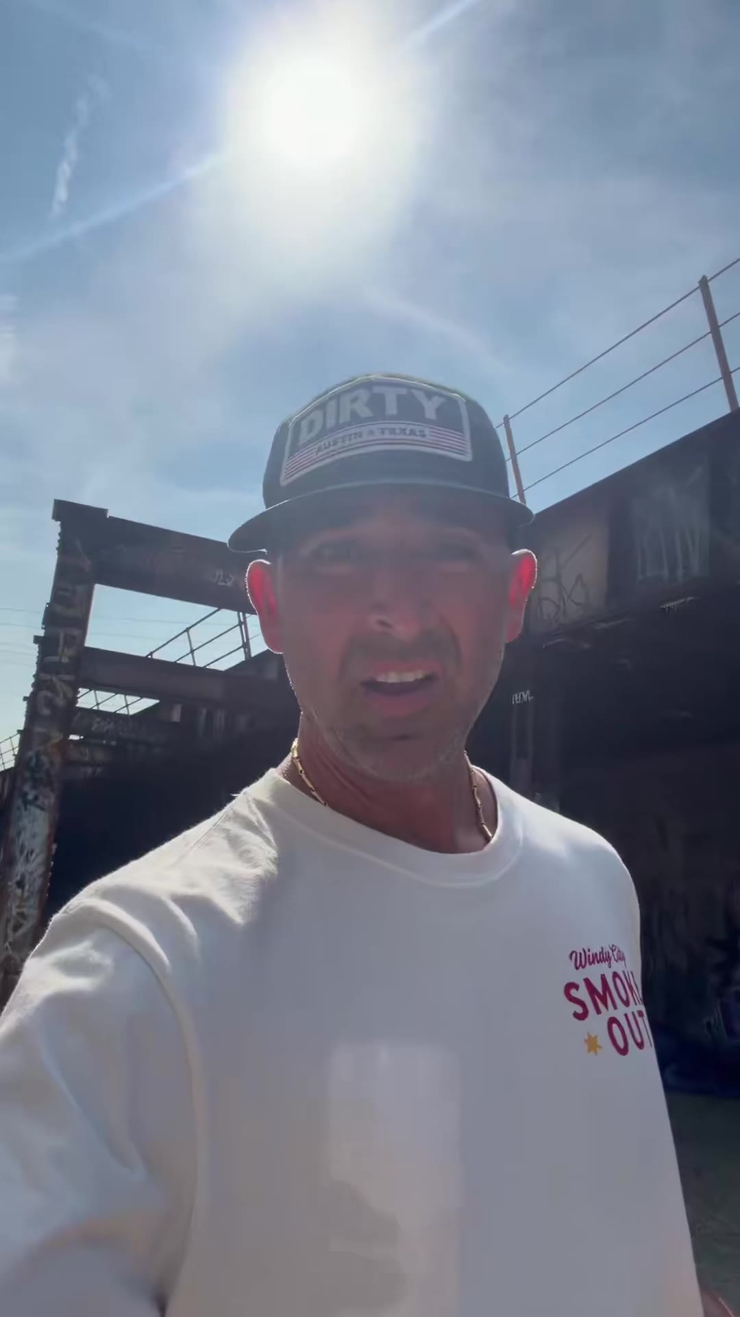 A man in a black "DIRTY Austin Texas" trucker cap and white "Windy City Smoke Out" t-shirt selfie-films himself walking through a graffiti-covered alley beneath a rusted iron railroad bridge, at one point pointing up at the bridge structure.