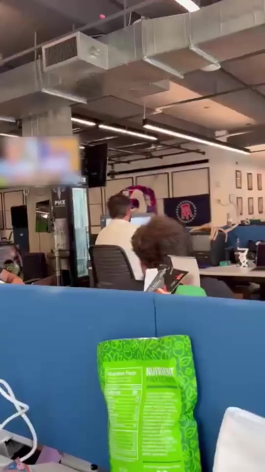 People in an office setting sit and stand near cubicles, with a TV screen showing blurred/pixelated content mounted on the wall; the space has industrial ceiling ductwork, a green protein/nutrition bag visible on a cubicle wall, and various office equipment.