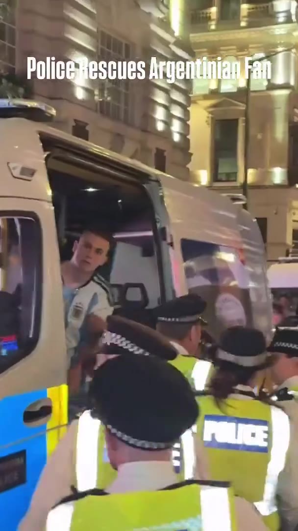 A crowd of soccer fans gathers at night in a European city street outside a Nike storefront, with a police officer in a hi-vis vest present amid the crowd.