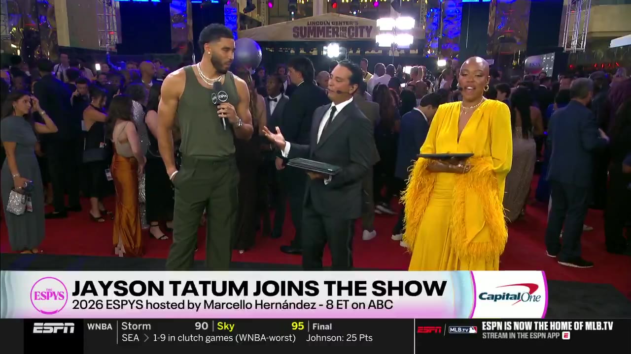 Jayson Tatum in an olive green tank top being interviewed on the red carpet at the ESPYs by a man in a suit and a woman in a yellow feathered dress, with a building and event crowd in the background.
