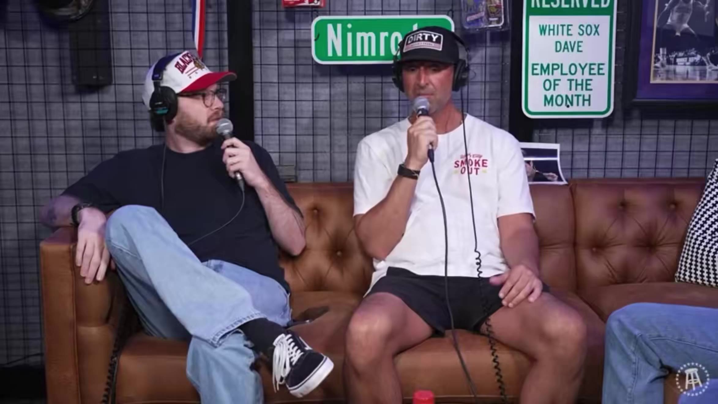 Two men sit on a leather couch in a podcast studio holding microphones, one wearing a red baseball cap, black t-shirt and jeans, the other wearing a dark cap, white t-shirt reading SMOKE OUT, and black shorts; the man in the cap briefly holds up a phone.