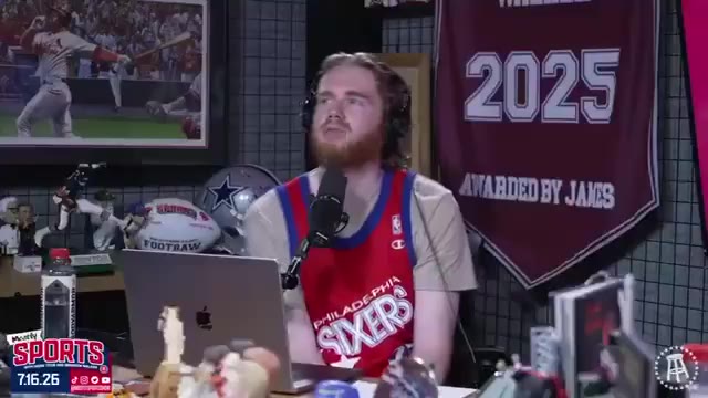 A man wearing a headset and USA-branded baseball-style jersey talks and gestures while seated at a podcast desk surrounded by sports memorabilia, bobbleheads, and merchandise.