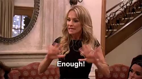 A blonde woman in a black top stands in a living room with a staircase and ornate mirror behind her, gesturing with both palms open while a caption reads 'Enough!'