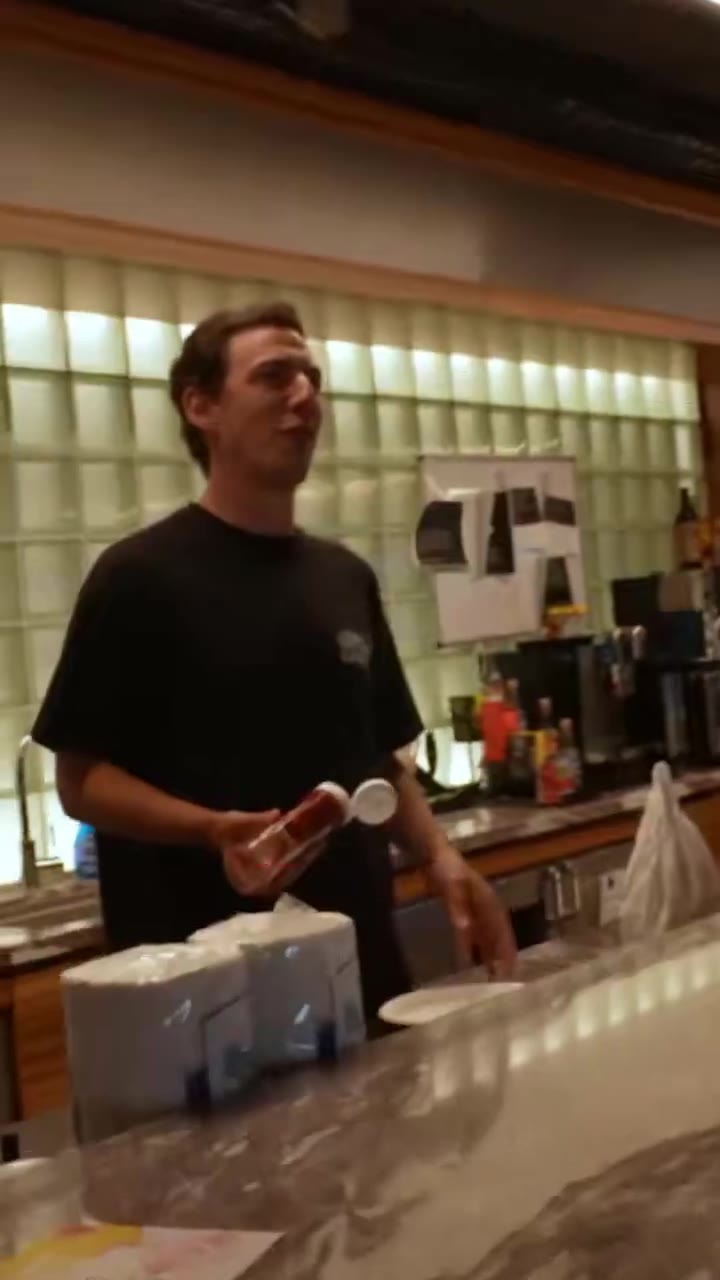 A man in a green t-shirt and cap sits in a wheelchair, one leg extended and bandaged, being pushed by another man in a bowling alley/lounge area with a golf simulator screen and stage with a drum kit visible in the background; earlier he stands in an office kitchen area holding a bag while talking.