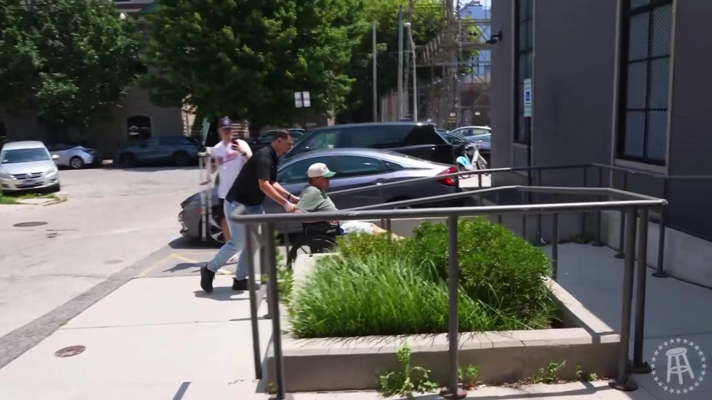 A man in a green shirt and cap is wheeled in a wheelchair by another man down an outdoor accessible ramp and into a building lobby with a hallway and a potted cactus visible.