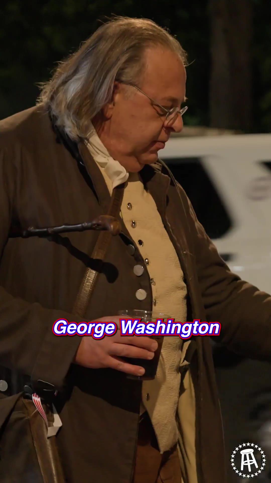A man in a colonial-era blue military costume rides a white horse down a street at night, waving to a crowd of onlookers as he passes a group of people including a man in a Benjamin Franklin costume.