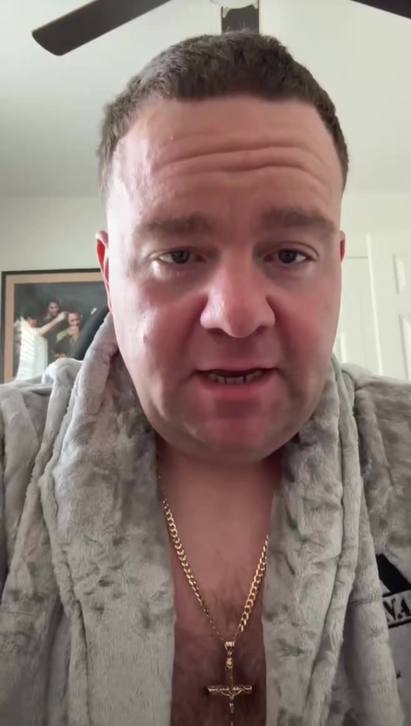 A man in a gray fur-collared robe with an open chest revealing a gold cross necklace and chain talks directly into a selfie camera in a bedroom, gesturing with an animated, wide-eyed expression, with a ceiling fan and framed picture visible behind him.