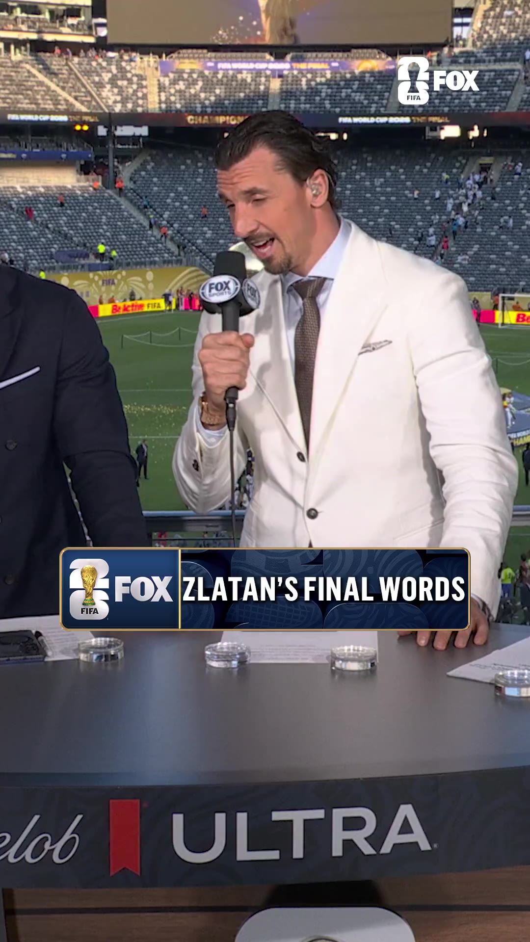 Zlatan Ibrahimovic, wearing a white suit, speaks into a FOX Sports microphone at a studio desk overlooking an emptying World Cup Final stadium, gesturing with his hands as he talks.