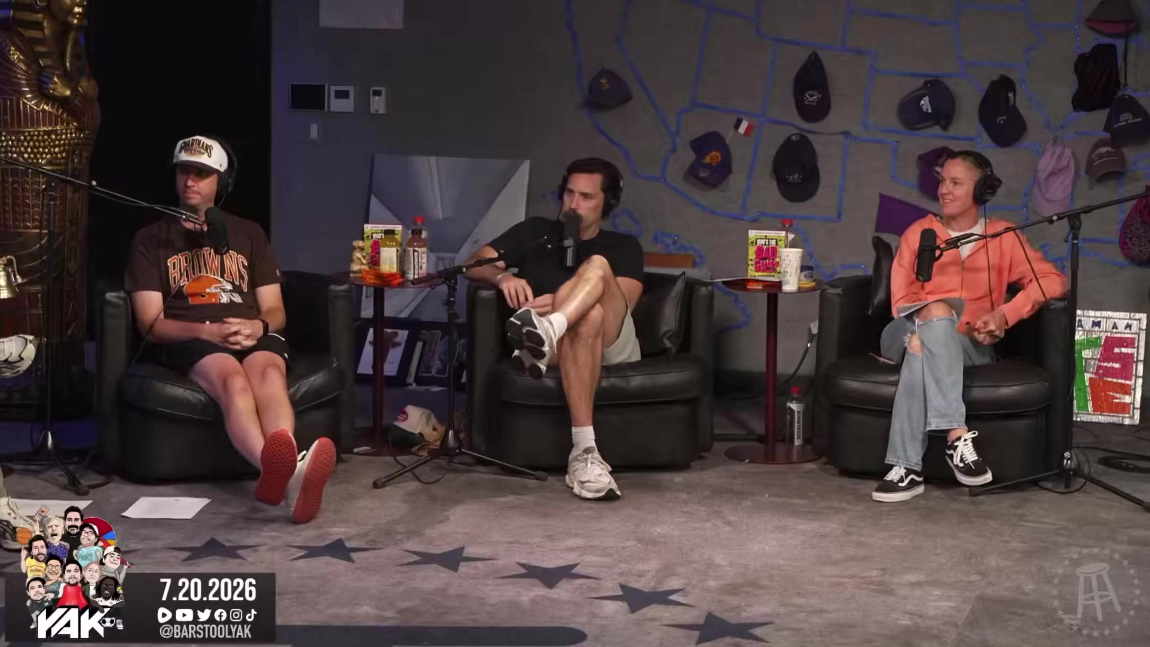 Three men sit in armchairs recording a podcast on a set decorated with a US map covered in baseball caps; the man on the left in a Cleveland Browns jersey and white cap gestures while talking, the man in the middle wears a black t-shirt and white sneakers, and the woman on the right in an orange top and headphones listens; a date overlay reads 7.20.2026 and a Barstool Yak logo/handle is shown.