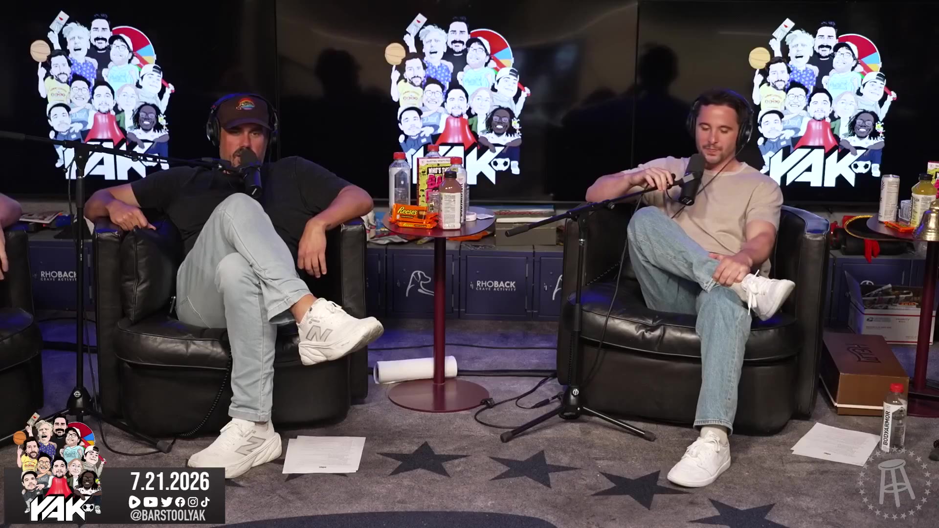 Two men sit in podcast chairs on a talk-show set with a mascot-collage backdrop and a monitor showing a Barstool Sports (PardonMyTake) tweet: Big Cat waving on a stadium video board at Wrigley Field with a Wintrust sign, captioned about him receiving a '50-50 big check from the Cubs.'