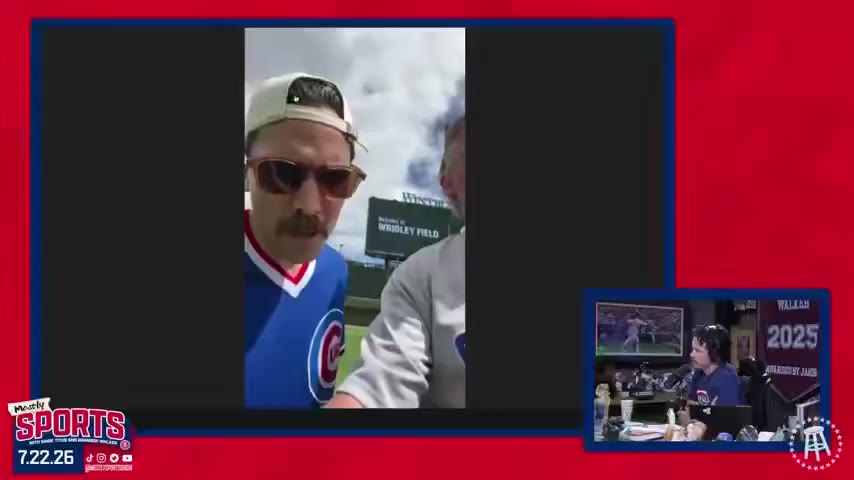 Two men at Wrigley Field, one wearing a Chicago Cubs pinstripe jersey and the other in a Cubs cap and sunglasses, filming a selfie video with the outfield and stands visible behind them.