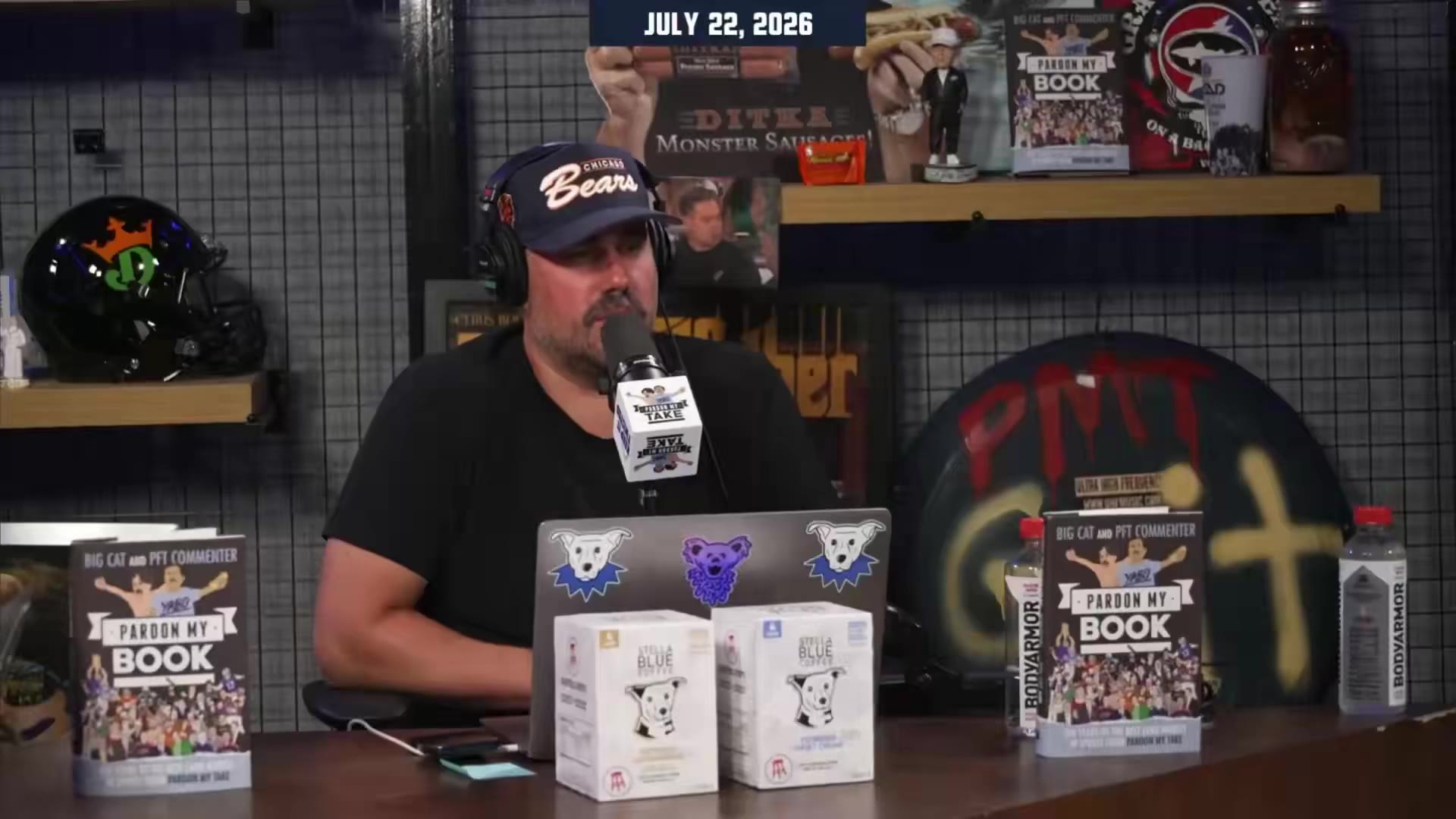 Two men in a podcast studio; one wearing a cap, headphones, and white t-shirt sits on a black leather couch pointing off-camera and speaking into a microphone, while a second man stands partially visible behind the couch in the background. On-screen date overlay reads JULY 22, 2026.