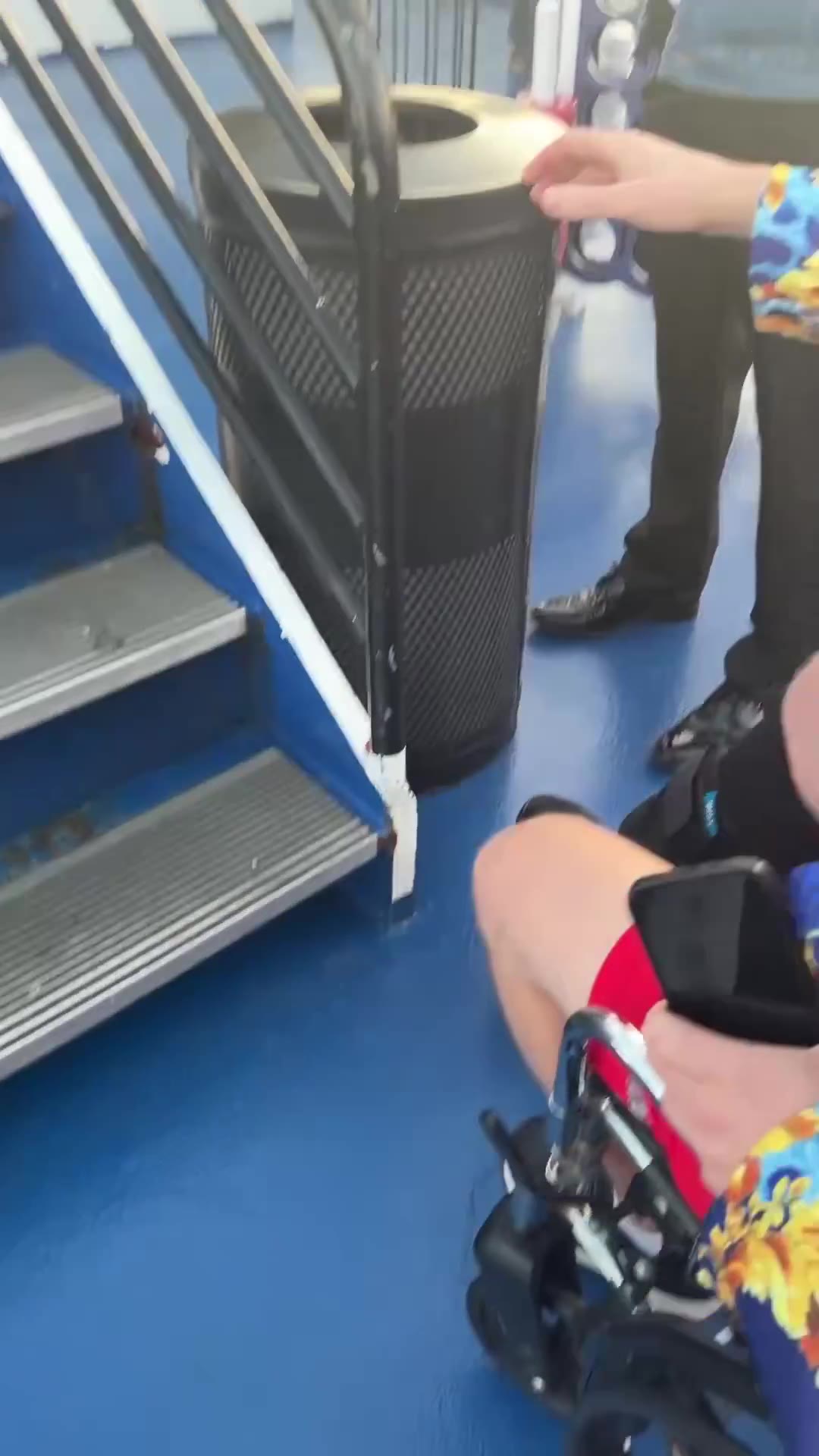 A person in a colorful patterned jacket and a leg brace walks up outdoor stairs on a boat deck, then is later seen seated in a wheelchair being pushed on deck near lounge chairs, a life preserver reading JASON STATHAM, and a movie poster for MUTINY, with a city skyline and water visible at sunset.