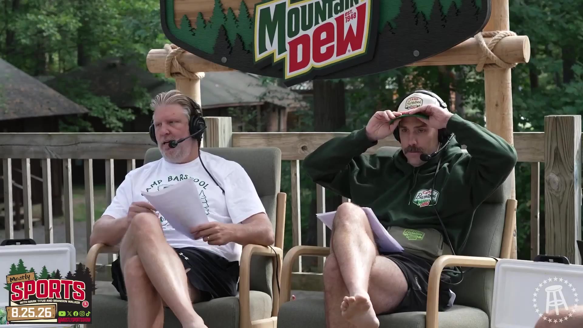 Two men sit in outdoor armchairs on a wooden deck under a Mountain Dew sign, wearing headset microphones; one in a white 'Camp Barstool' t-shirt gestures with his hands raised while talking, the other in a green Barstool hoodie and cap holds papers and gestures back.
