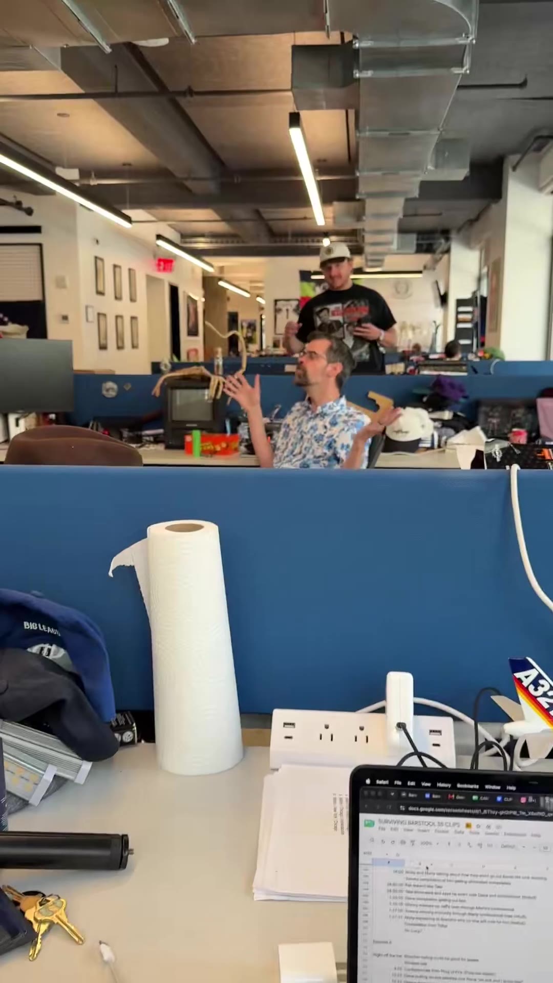 A man with glasses and a graying beard, wearing a blue floral shirt, sits at an office desk cluttered with baseball caps, snack boxes, and a Sylvania TV, gesturing with his hands while talking; office cubicles, a Barstool jersey, and a flag are visible in the background.