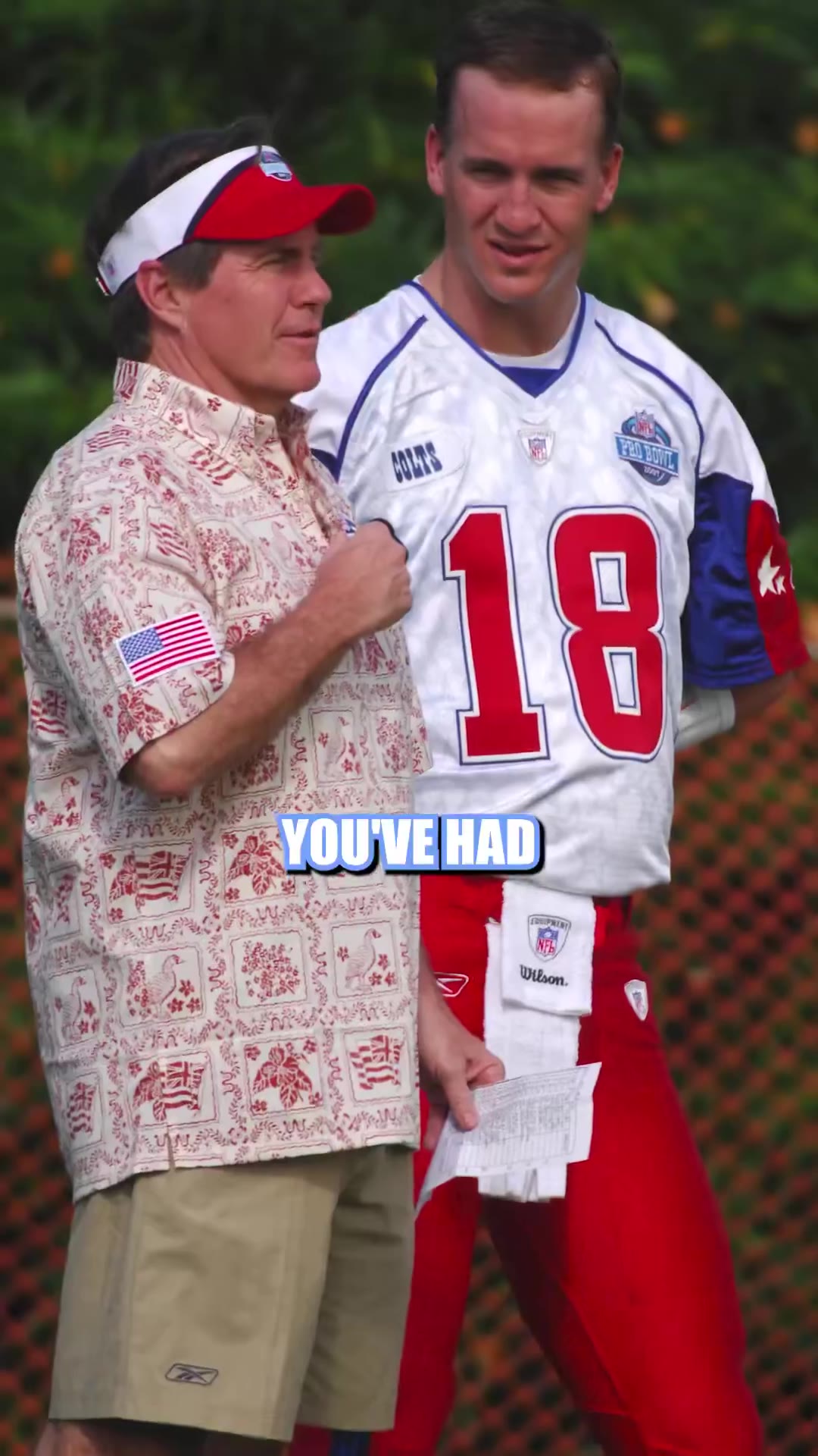 Two men seated in armchairs in a sports-memorabilia-filled studio talk on a podcast; one man wears a cap and t-shirt, the other, an older man in a dark quarter-zip and shorts, gestures while speaking near a display case of championship rings and a Super Bowl trophy replica.