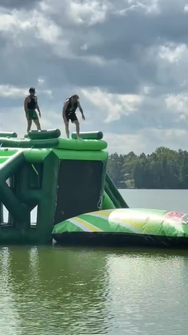 A shirtless man in green shorts stands atop a large green inflatable water obstacle course floating on a lake, then jumps down onto a smaller inflatable 'blob' pillow, causing a big splash while another person swims nearby in the water.