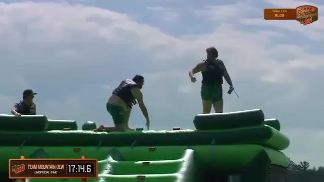 A person swims/wades through choppy water after an inflatable obstacle course feature launches them into the water; an on-screen graphic reads 'Team Mountain Dew 17:21.7 Unofficial Time'.