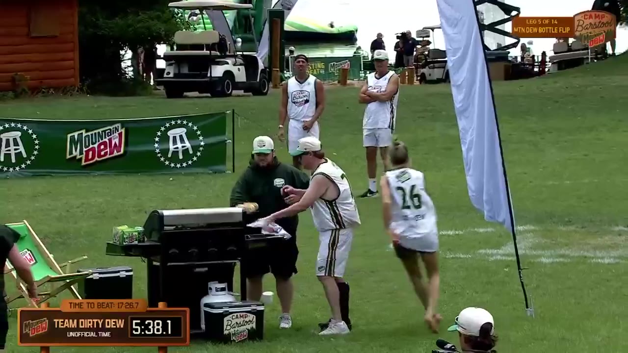 A man in a green-and-white Camp Barstool tank top eats a hot dog while being interviewed with a microphone reading a competitor time (6:04.0 to 7:06.4), on-screen graphics show 'LEG 6 OF 14, GLIZZY GOBBLE' and 'TEAM DIRTY DEW' with 'Time to Beat: 17:26.7', in an outdoor camp setting with tents and a wooded backdrop.