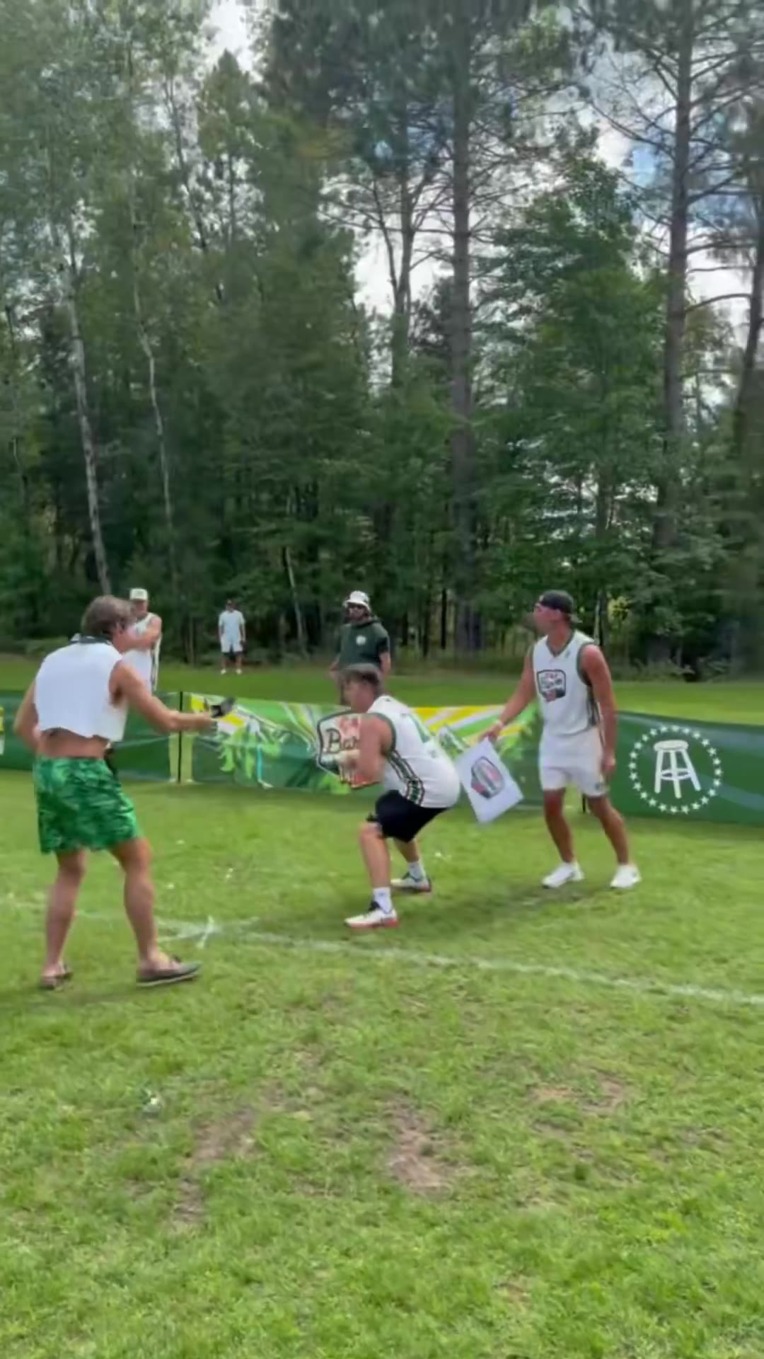 Two men in green Camp Barstool staff shirts wrestle a shirtless man in green shorts to the ground on a grass field, then the shirtless man walks off with them near a Camp Barstool banner.