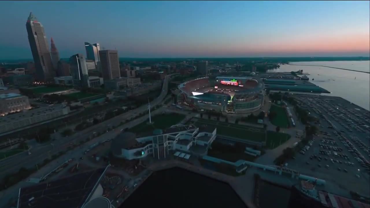 Aerial drone shot of downtown Cleveland skyline and Lake Erie with Cleveland Browns Stadium lit up at dusk, followed by broadcast footage of a Cleveland Browns kickoff coverage team running downfield against the New England Patriots, on-screen scoreboard reading Patriots 0, Browns 7, 1st quarter, 11:39, Thursday Night Football.