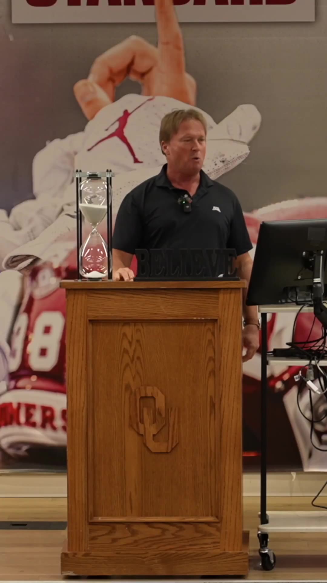 Jon Gruden speaking to a seated audience in a lecture hall with a projector screen reading 'WHAT LIES AHEAD - PHIL 3:13' behind him, standing near a podium branded with an Oklahoma football logo.