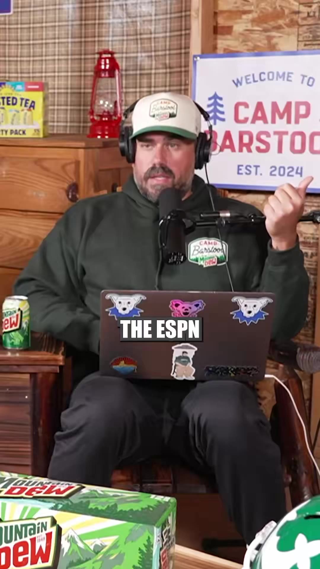 Two men wearing Camp Barstool trucker hats and headsets sit in a rustic cabin-style podcast studio talking into microphones with laptops on their laps, one in a green hoodie and one in a white polo and sunglasses with a green football helmet on the table.