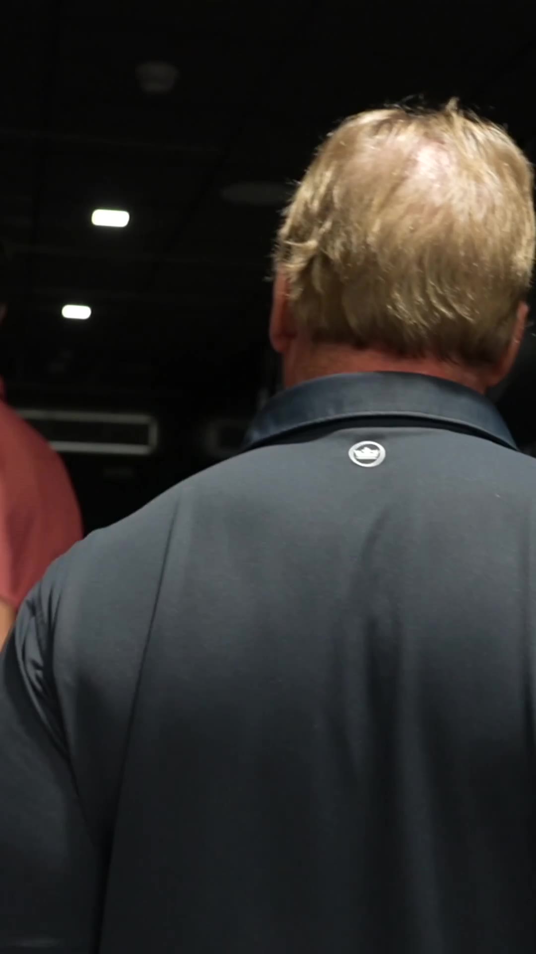 A man in a dark jacket walks out of a stadium tunnel into daylight, then stands looking over an empty stadium bowl with maroon and gray seating, a video board reading partly 'OU' with a Jordan brand logo, and a running track/football field below.