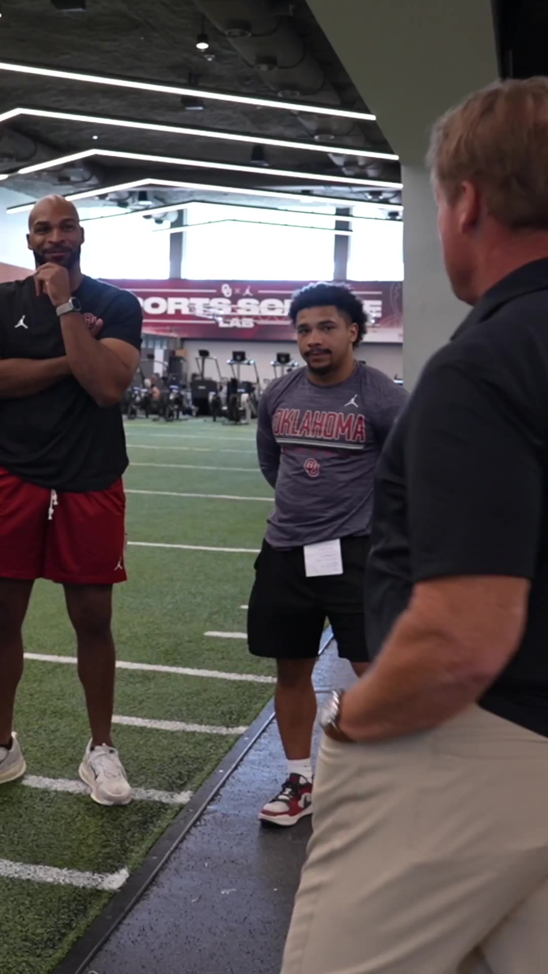 A shirtless man in a cap and black shirt stands on an indoor turf football field; a bald man in a black 'Built By Oklahoma' shirt and red pants talks with a man in a black polo and khakis with his back to the camera, then several men including one in an Oklahoma-branded black shirt gather and smile together on the field.