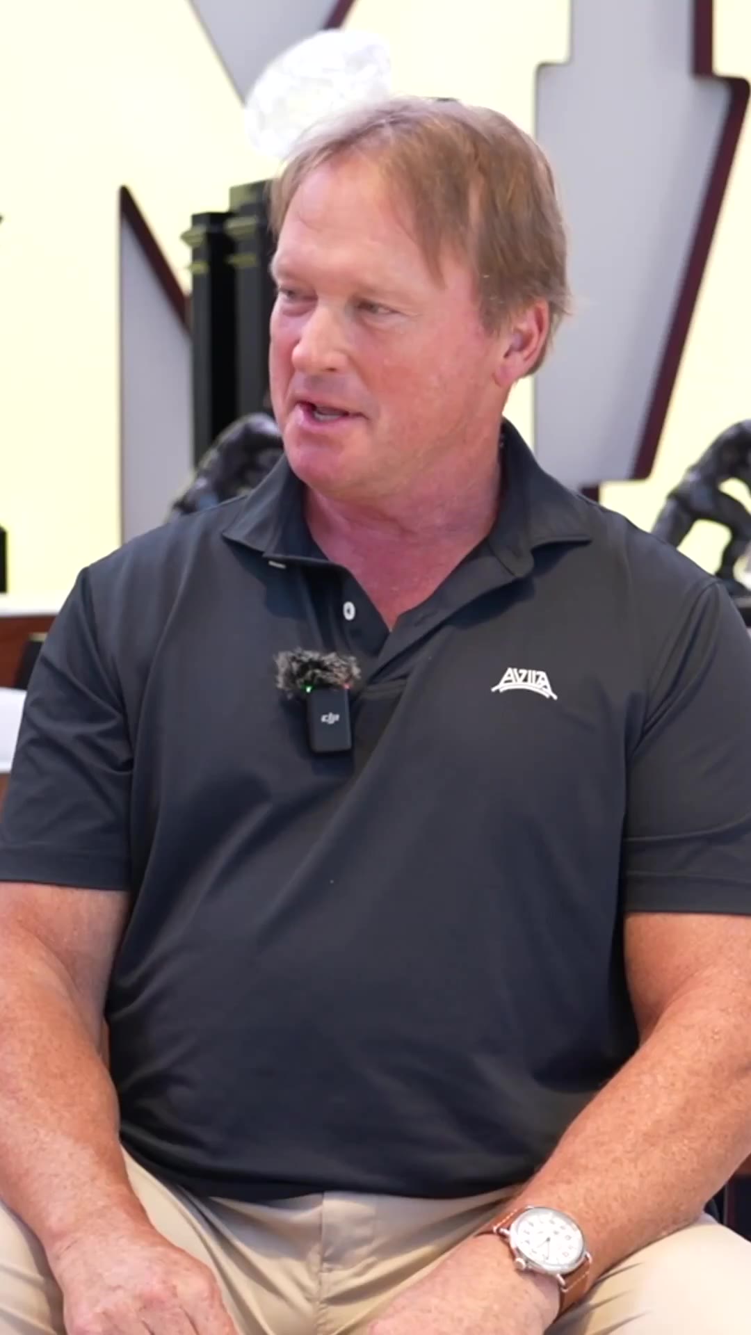 A young man with curly brown hair wearing a red University of Oklahoma t-shirt sits in a chair being interviewed indoors, smiling and laughing, with an OU football trophy case and clip-on lavalier microphone visible in the background.