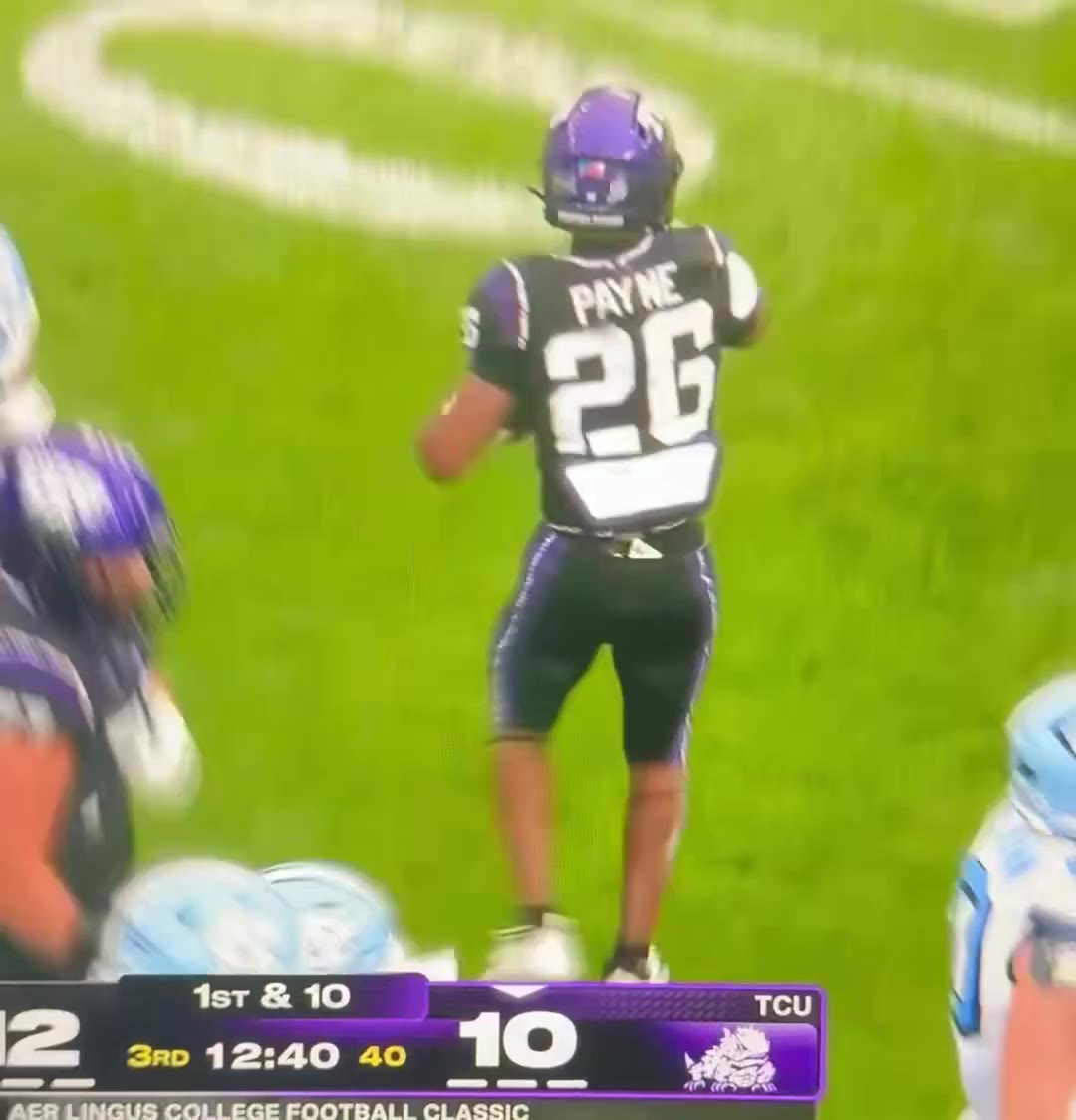 TCU defensive player number 26, jersey name PAYNE, raises his arm and celebrates near midfield after a play against North Carolina, with a referee's yellow/red flag markers visible on the field; scoreboard shows North Carolina 12, TCU 10, 3rd quarter 12:37-12:39.
