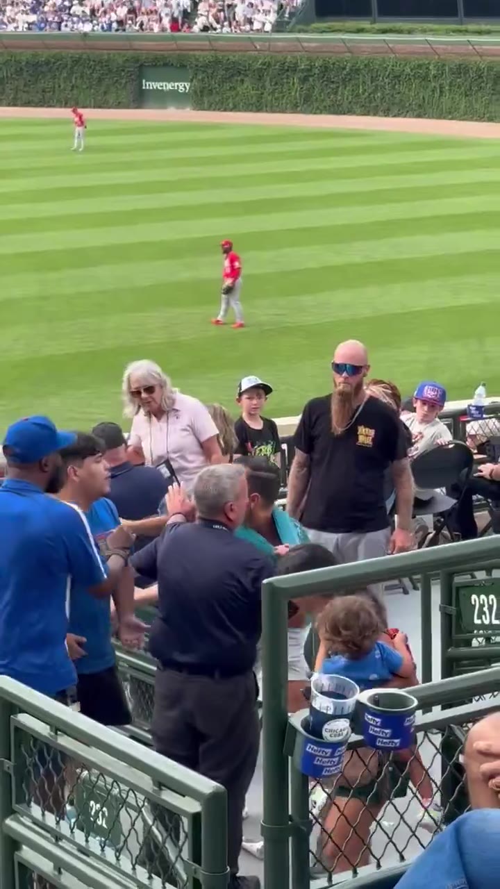 Fans in the outfield bleacher seats at Wrigley Field turn and react excitedly, with some climbing over seats and railings while others look surprised or point, as a ball appears to land nearby among the crowd; the stadium scoreboard and jumbotron are visible in the background.