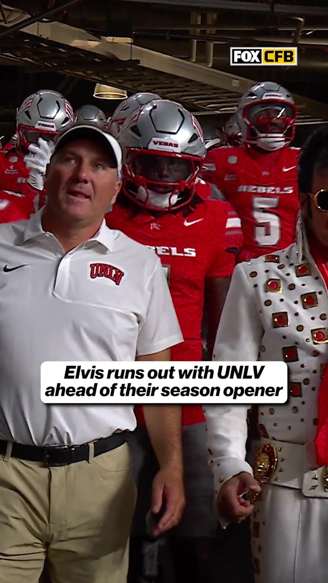 A man dressed as Elvis Presley in a white jumpsuit and red scarf runs out onto the field alongside UNLV Rebels football players in red jerseys and silver helmets, past a coach in a white polo and visor, inside a stadium with a UNLV scoreboard visible.