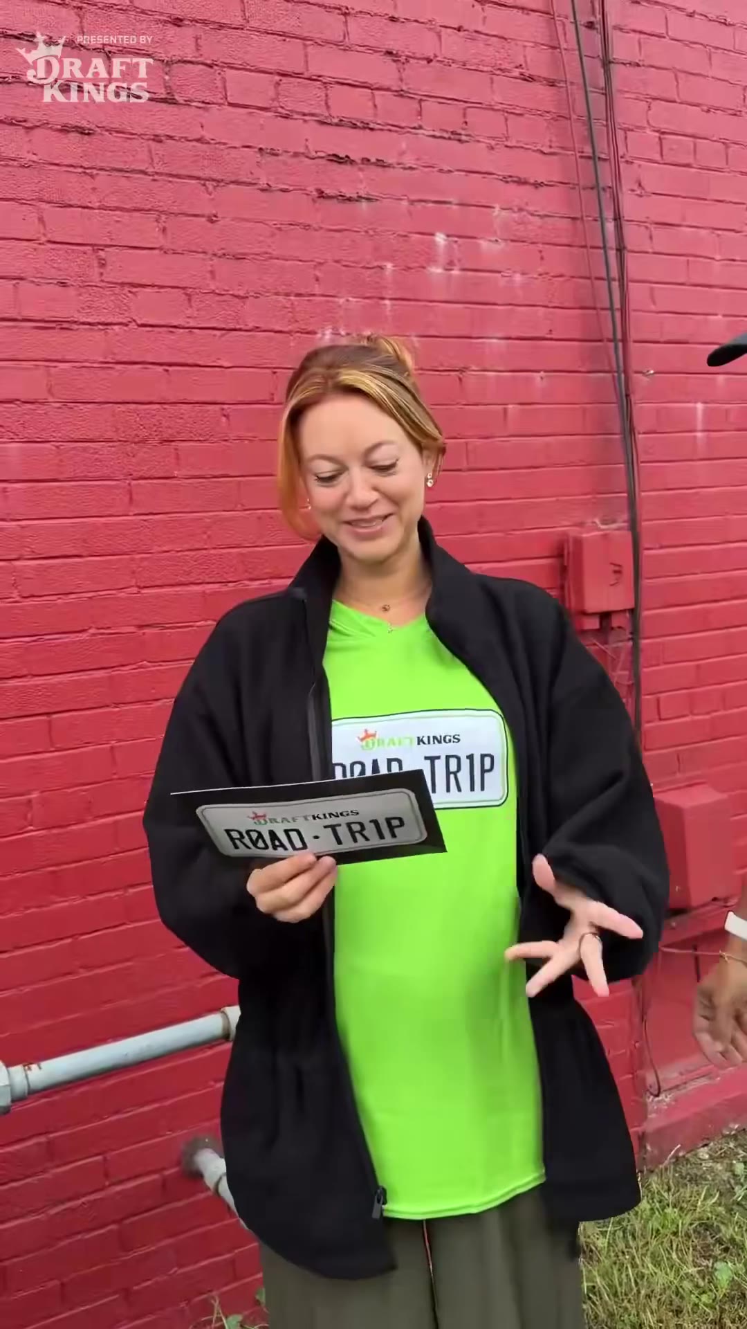 A woman in a green DraftKings road-trip shirt sits at a bar table with a large paper cone of breaded buffalo wings/cheese curds and several dipping sauces, laughing and reading a card outside a brick building beforehand.