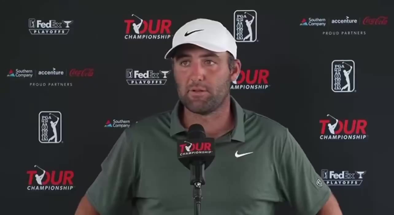 A man in a white golf cap and dark green polo shirt speaks at a podium microphone in front of a Tour Championship / FedEx Cup Playoffs step-and-repeat backdrop, looking down and away from the camera at times.