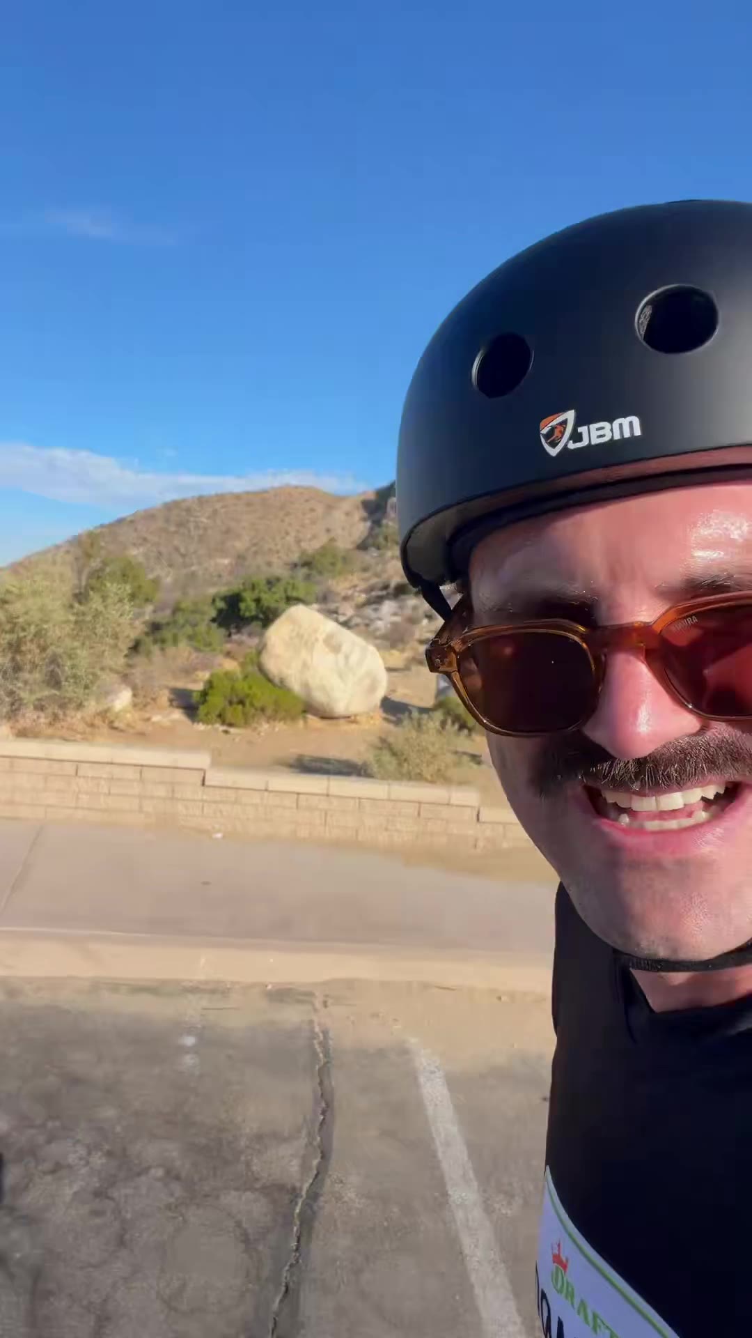 A person on rollerblades falls backward and lands sitting against a low stone/concrete wall on a paved path near a desert hillside, wearing a helmet, elbow pads, and knee pads, laughing while holding a phone; his shirt reads DraftKings Road Trip.