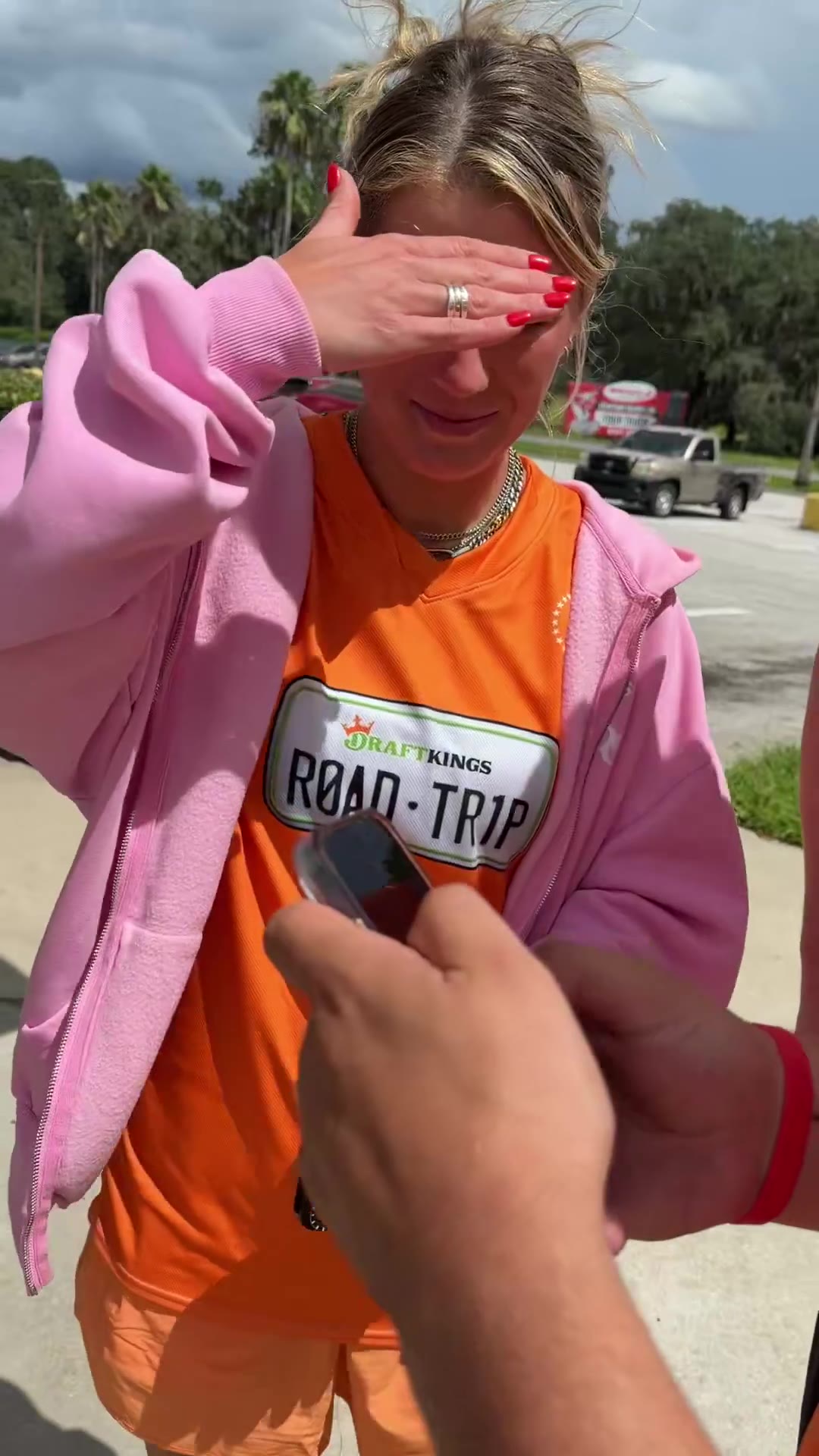A man in an orange DraftKings Road Trip t-shirt and backwards cap poses with a group outside, then takes a photo inside a store with another man wearing a Blue's Clues-style t-shirt and backwards blue cap, then stands alone outside checking his phone under a stormy sky near a gas station.