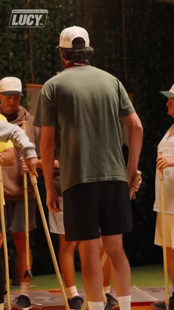 A group of men on a set with astroturf flooring hold long croquet-style mallets while standing near each other; one man wears a striped headband and a blue shirt, another an LUCY-branded overlay is visible in the corner; a later frame shows a close-up of a bearded man speaking to camera.