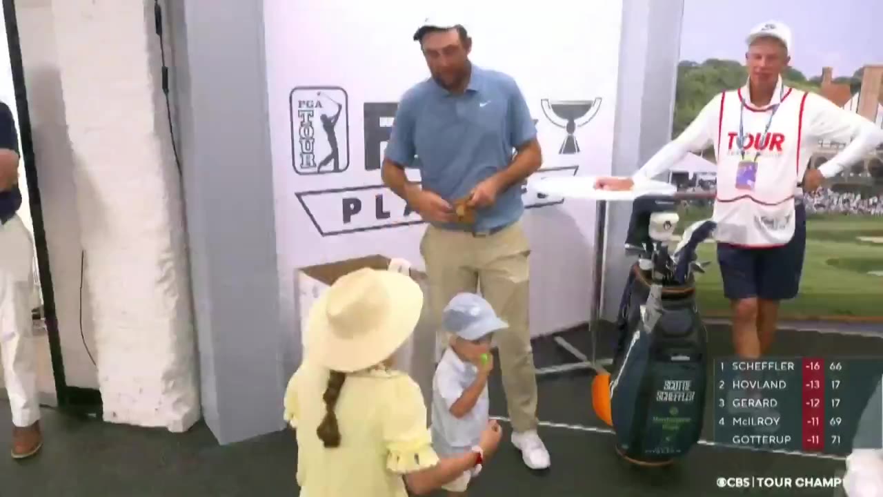 A golfer in a light blue polo interacts with young children, including a toddler in a matching blue hat and outfit, in a tented backstage area with a FedEx Cup Playoffs backdrop and golf bag reading 'Scheffler'; an on-screen leaderboard shows Scheffler leading at -16.