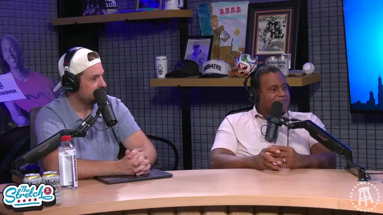 Two men seated at a podcast desk wearing headphones and holding microphones, one in a white cap and light blue polo gesturing with clasped hands, the other in a white textured polo gesturing with both hands raised while talking.