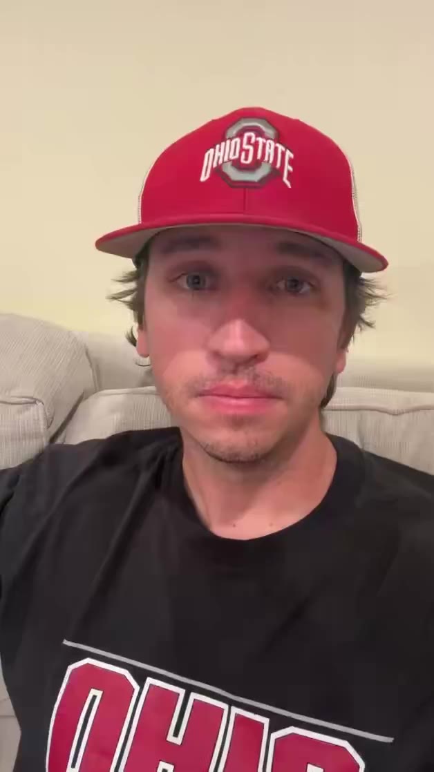 A man wearing an Ohio State cap and black Ohio State t-shirt speaks directly to the camera while seated on a couch, gesturing with his hands.